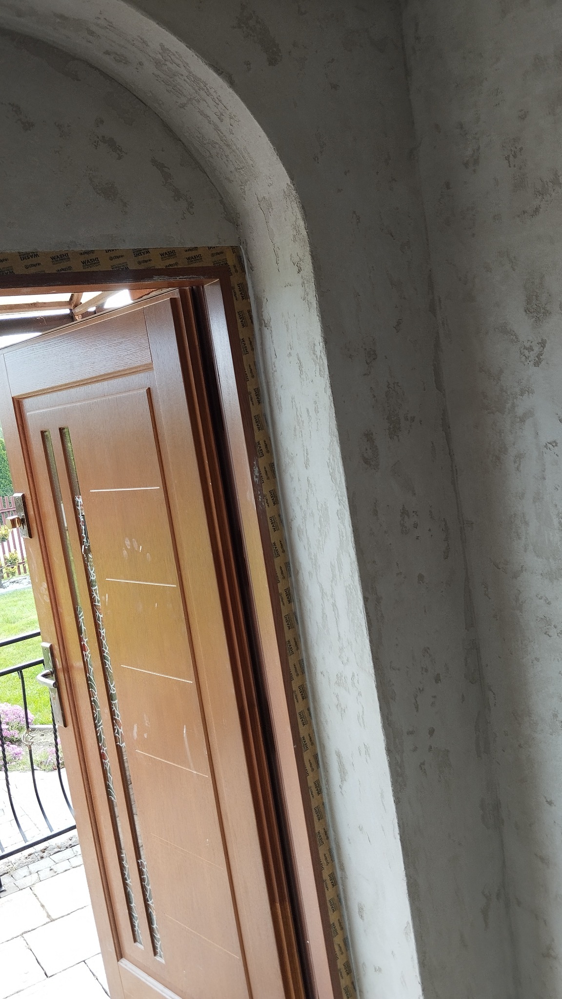 Otwarta, drewniana brama wejściowa z widocznymi śladami szpachlowania na łuku i ścianie. Widoczne zabezpieczenia taśmą malarską. Ujęcie z bliska, z zewnątrz.