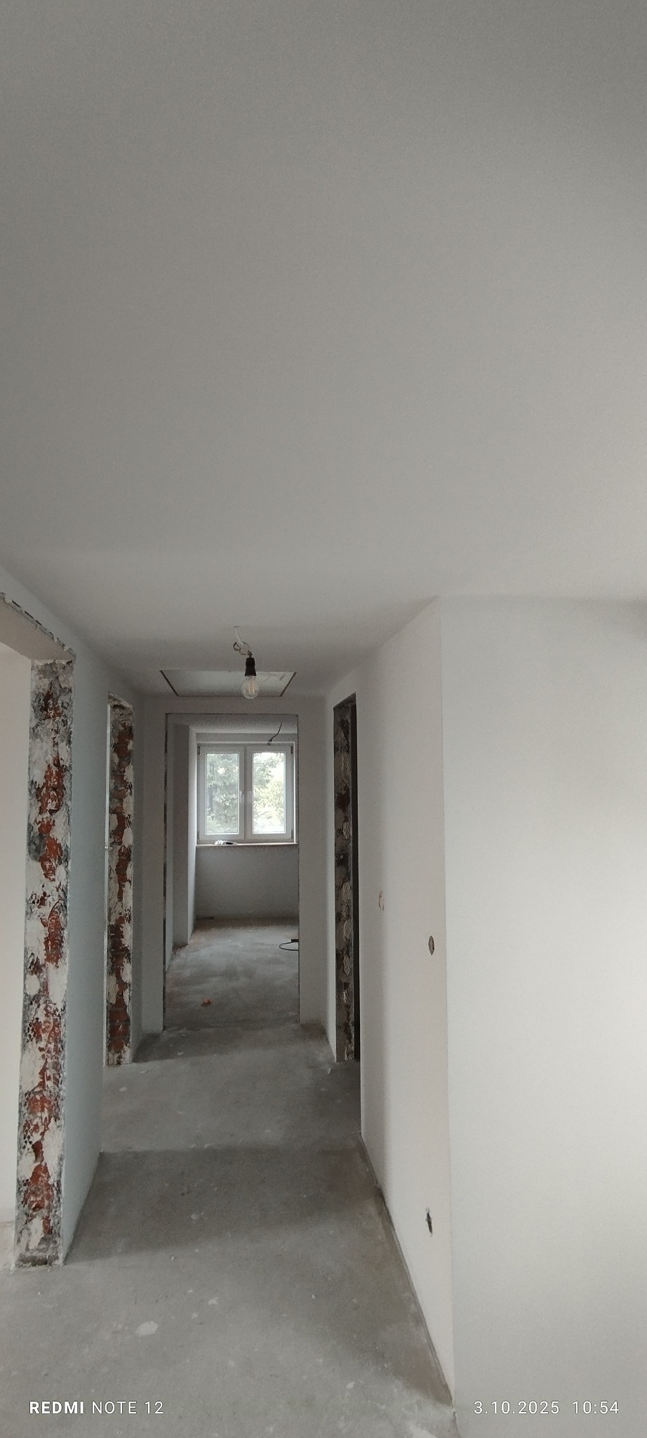 Korytarz w trakcie remontu: świeżo pomalowane ściany na biało, widoczne fragmenty odsłoniętej cegły przy futrynach, surowy beton na podłodze, okno w tle.