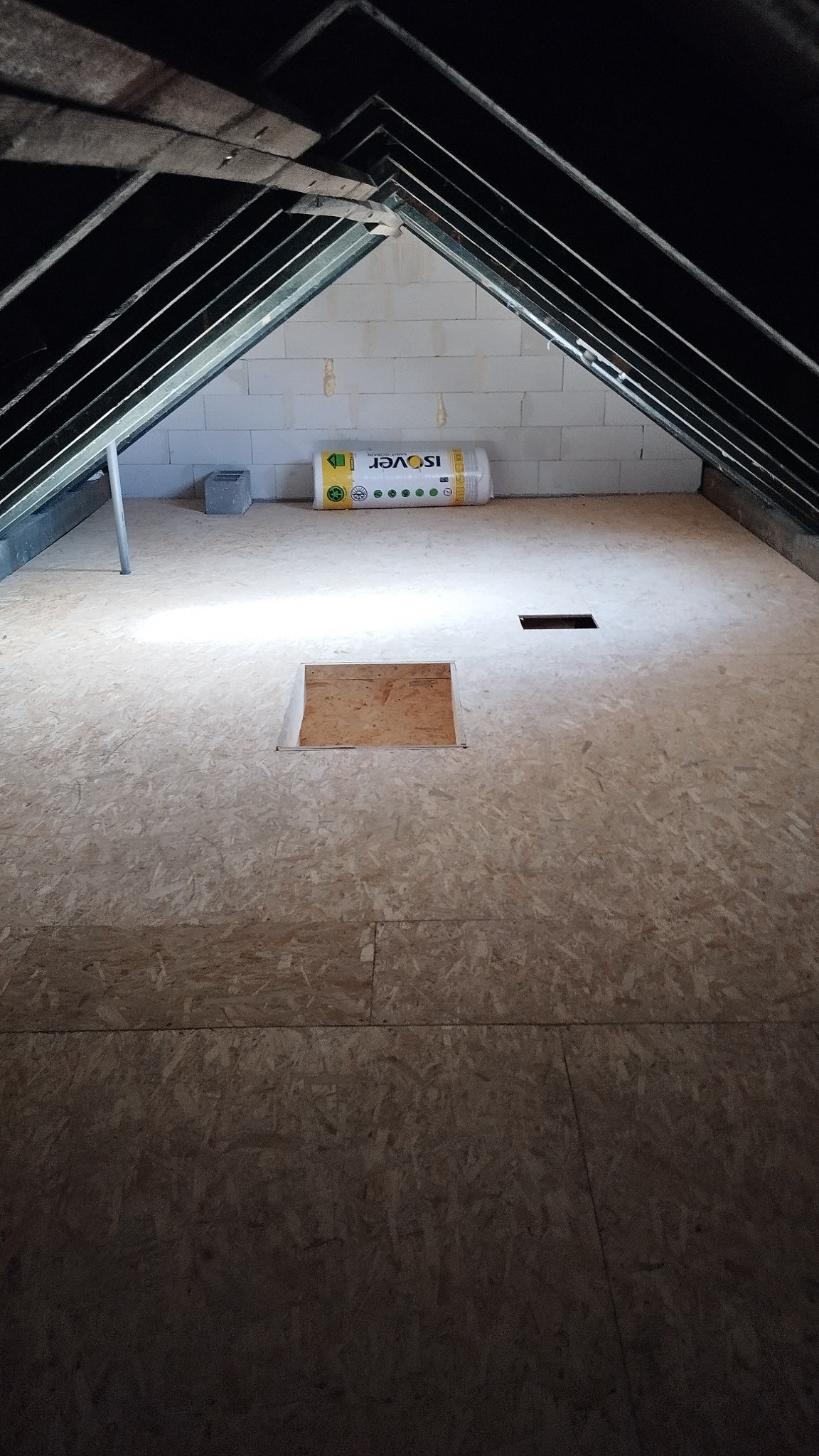 Poddasze z płyt OSB, widoczna izolacja Isover przy ścianie z bloczków. Otwór w podłodze i czarna kratka wentylacyjna. Metalowa konstrukcja dachu.