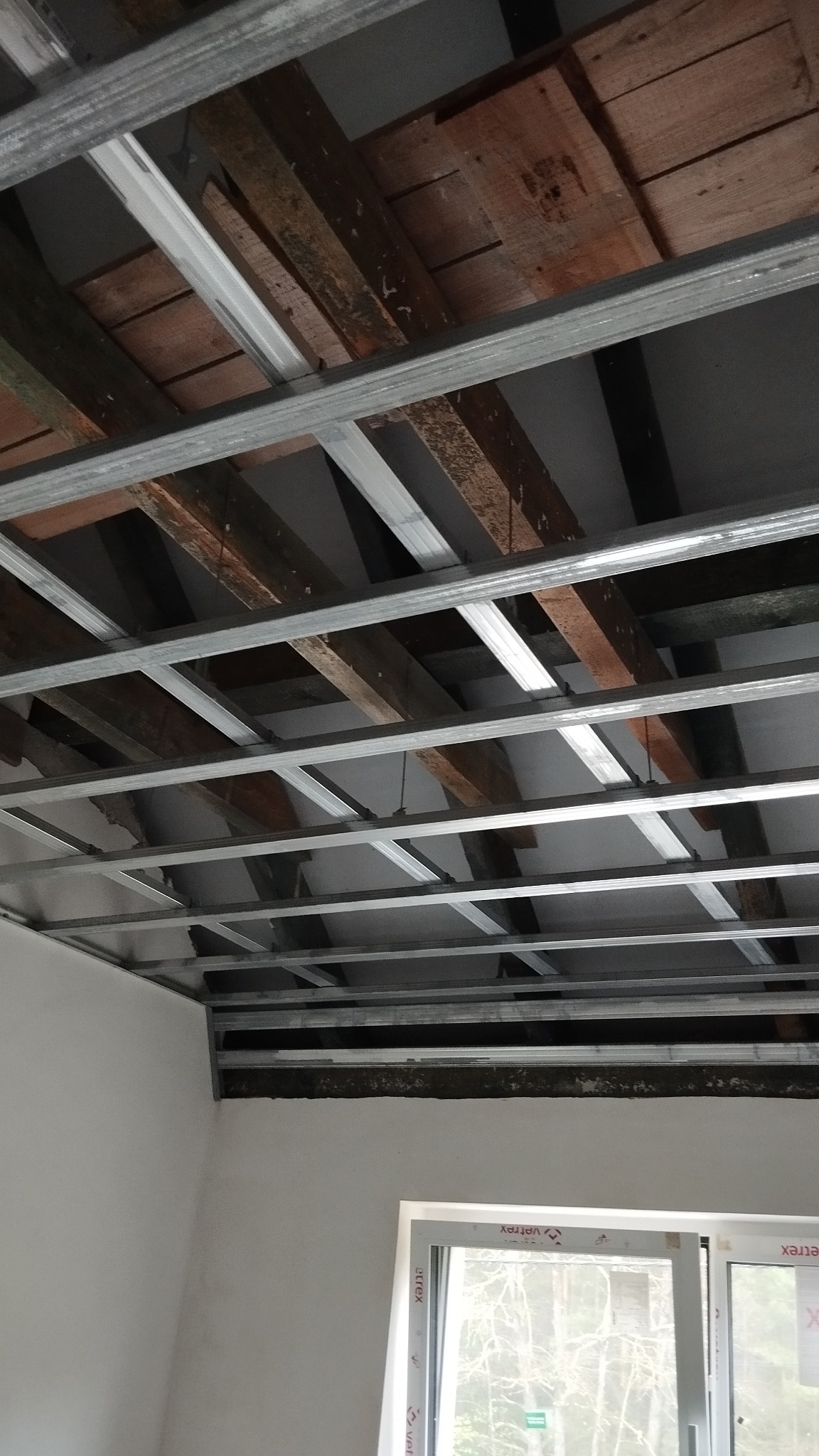 Metalowa konstrukcja sufitu podwieszanego z widocznymi drewnianymi elementami stropu, w tle okno z widokiem na zewnątrz. Prace wykończeniowe w pomieszczeniu.