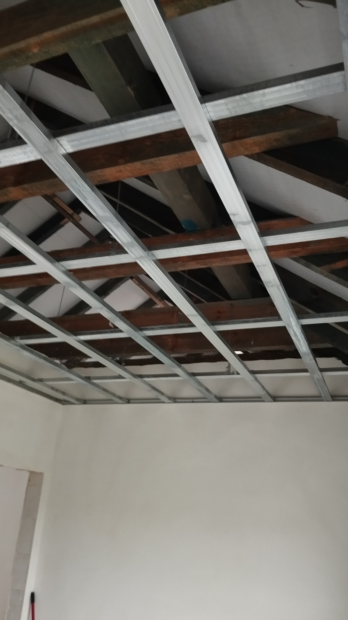 Montaż metalowej konstrukcji sufitu podwieszanego z widocznymi drewnianymi elementami więźby dachowej i fragmentem białej ściany.