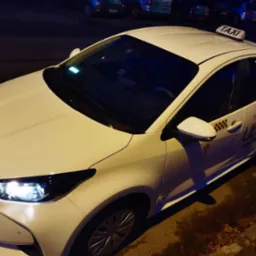 Biała Toyota Yaris z napisem TAXI na dachu i logo Uber na boku, zaparkowana na ulicy nocą.