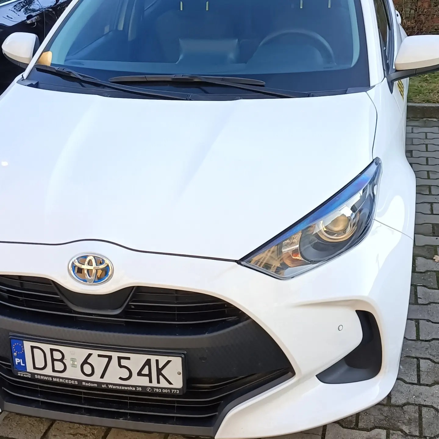 Biała Toyota Yaris z widoczną tablicą rejestracyjną DB 6754K na tle kostki brukowej.