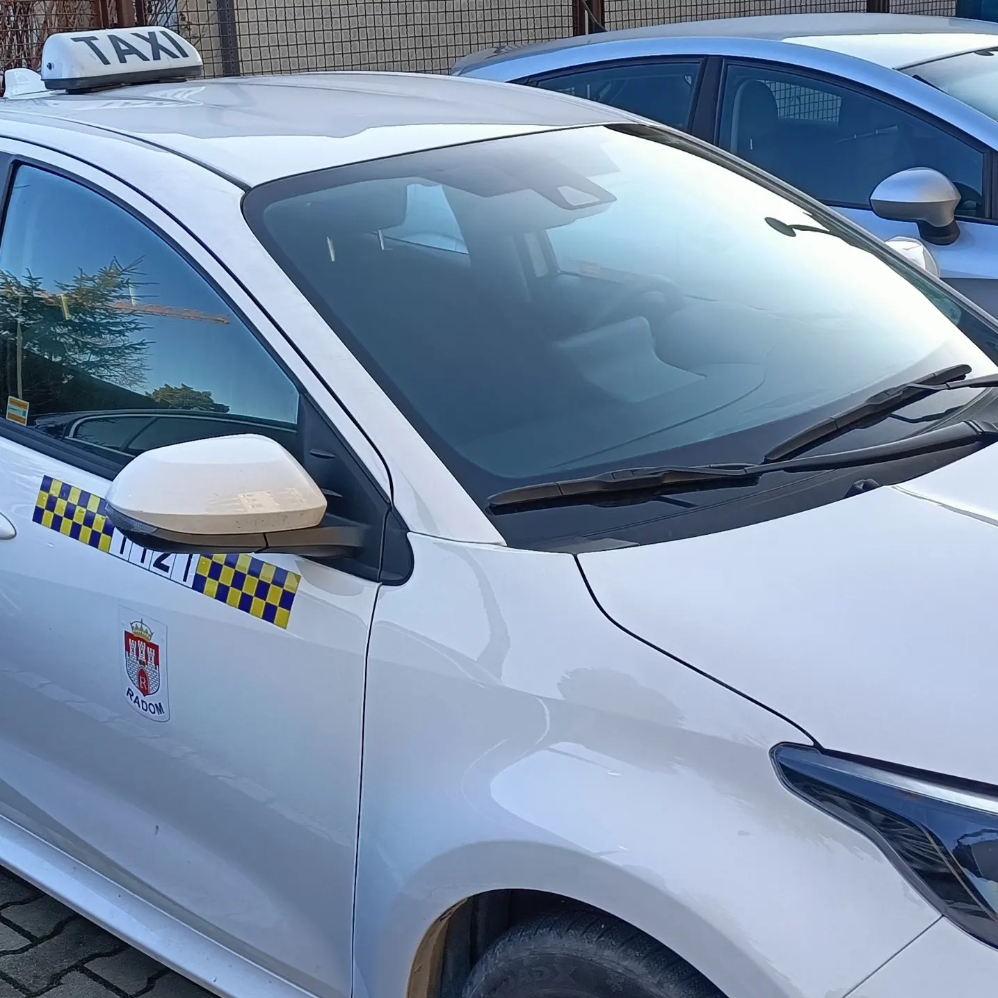Biały samochód taxi z logo Radomia na boku i charakterystyczną szachownicą, zaparkowany obok innego pojazdu.