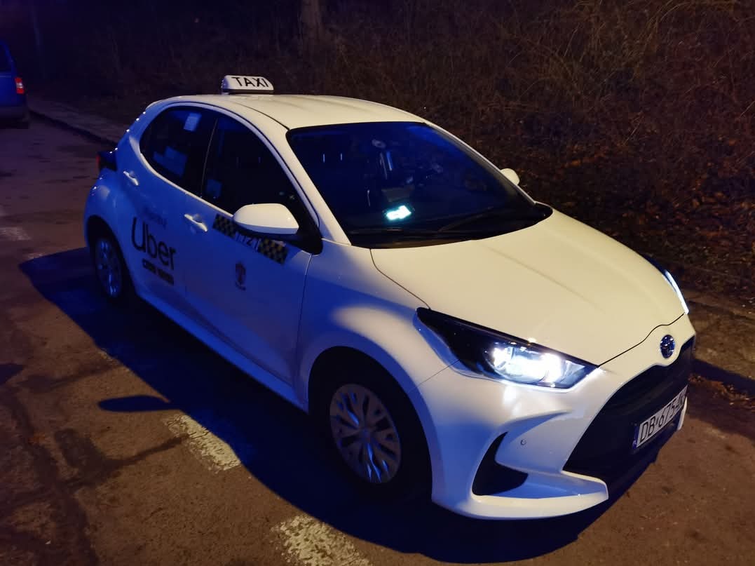 Biała Toyota Yaris z oznaczeniem Uber i TAXI na dachu, zaparkowana na ulicy w nocy, widok z przodu i boku.