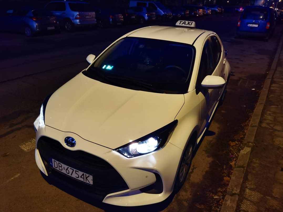 Biały samochód osobowy z napisem TAXI na dachu, zaparkowany na ulicy w nocy, widok z przodu, w tle inne pojazdy.