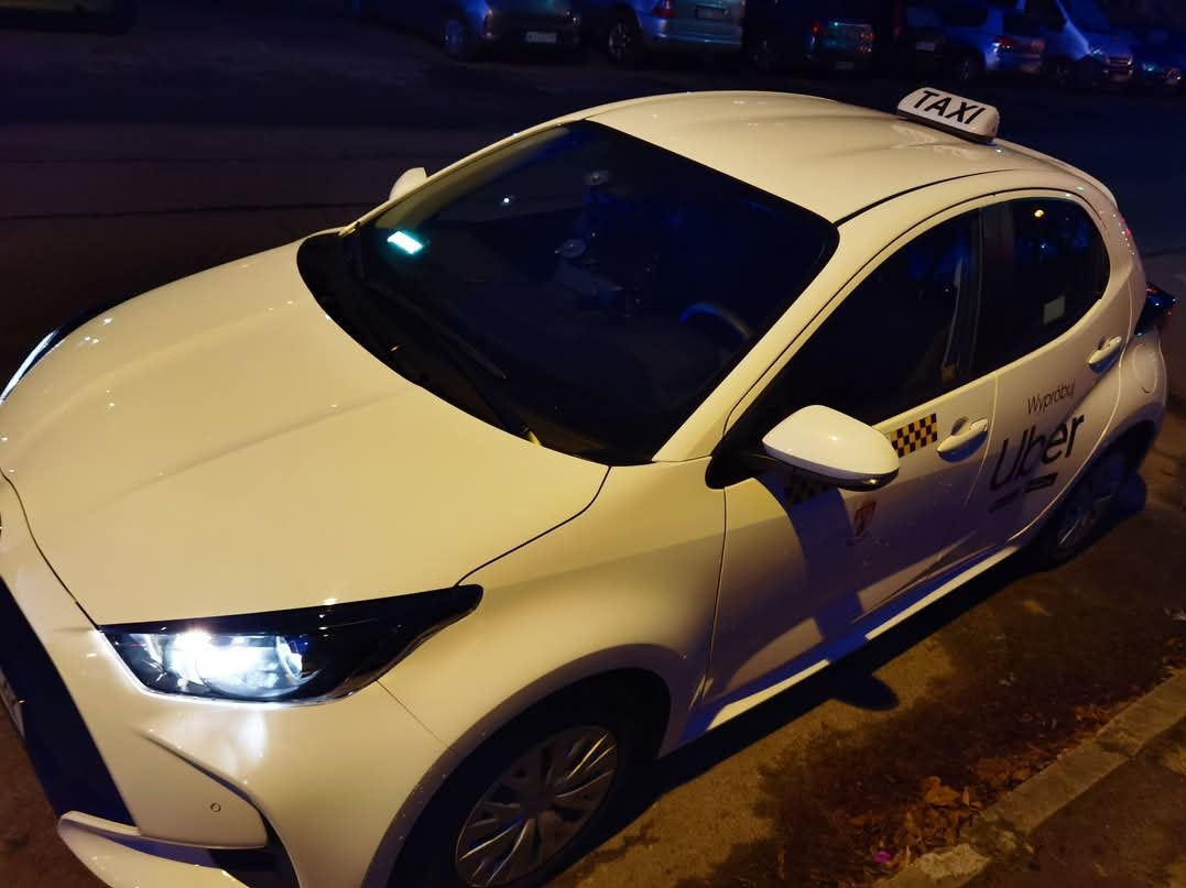 Biała Toyota Yaris z napisem TAXI na dachu i logo Uber na boku, zaparkowana na ulicy nocą.