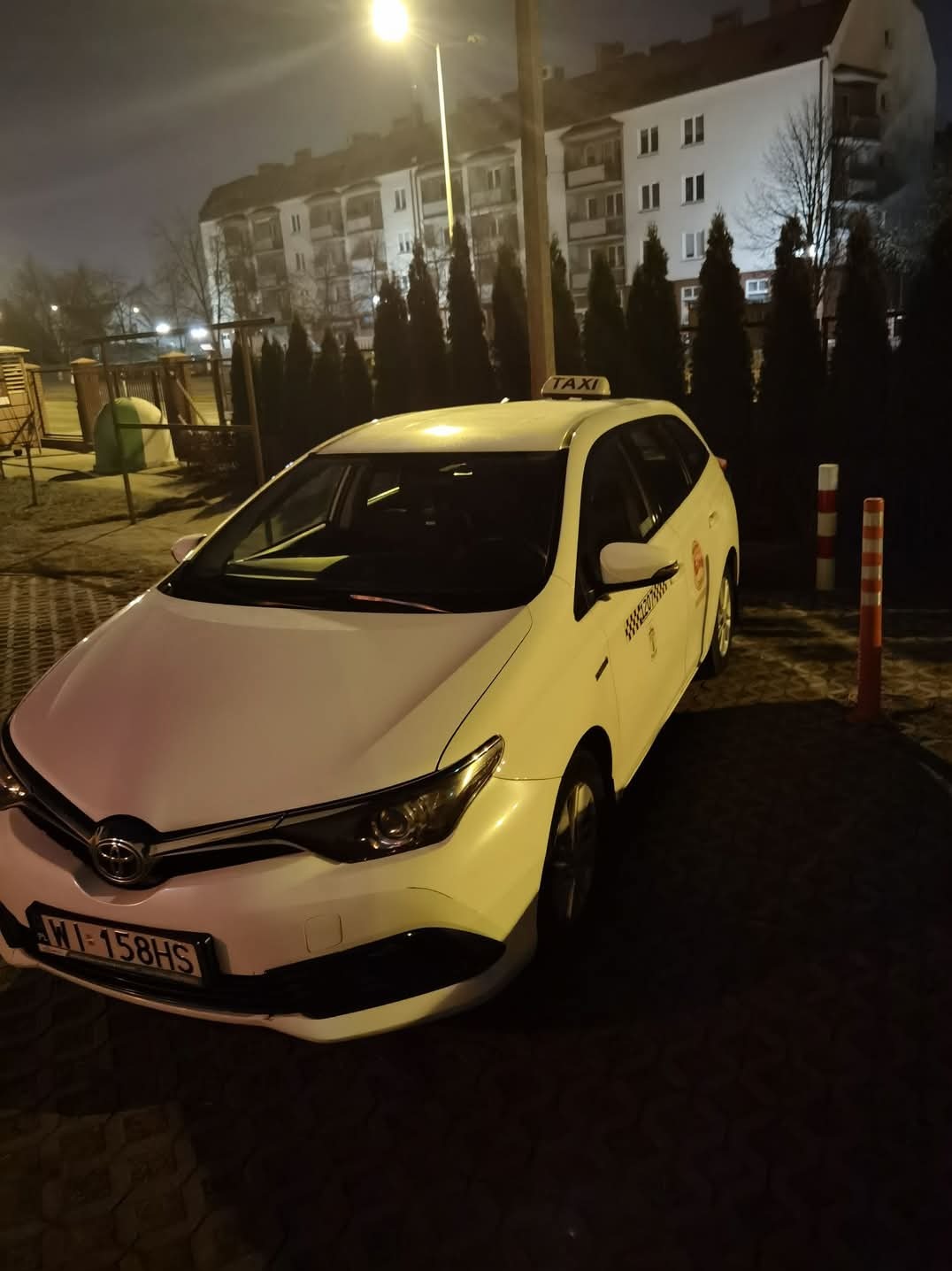 Biała taksówka kombi Toyota Auris zaparkowana nocą na tle bloku mieszkalnego i rzędu iglaków.