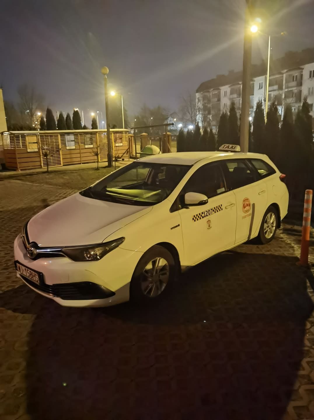 Biała taksówka kombi marki Toyota Auris z numerem bocznym i napisem 'Taxi' na dachu, sfotografowana nocą na brukowanym parkingu, w tle budynek mieszkalny i rzędy drzew.