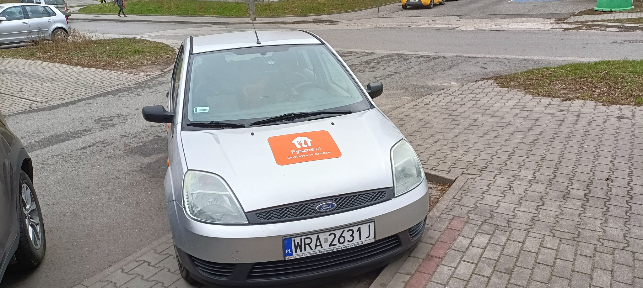 Srebrny Ford Fiesta z naklejką Pyszne.pl na masce, zaparkowany na chodniku z kostki brukowej.