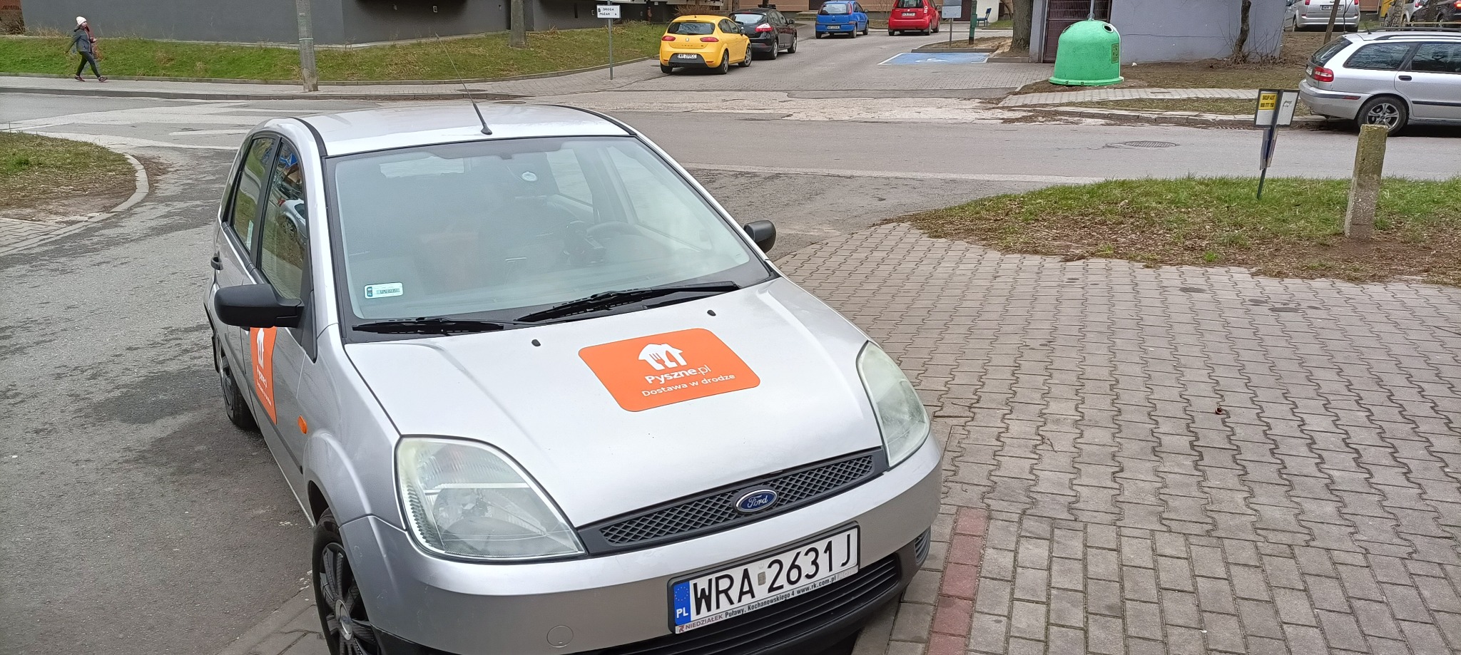Srebrny samochód z pomarańczowym logo Pyszne.pl na masce zaparkowany na chodniku z kostki brukowej.