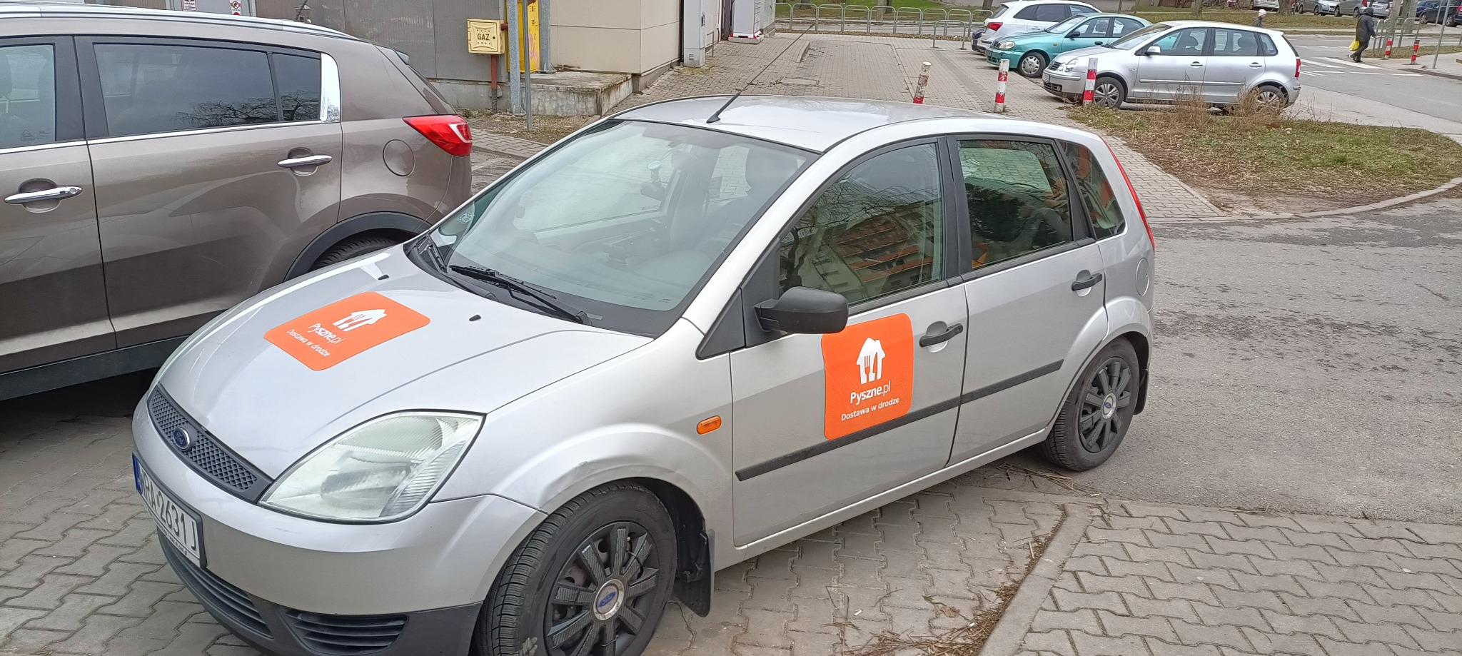 Srebrny Ford Fiesta z pomarańczowymi naklejkami Pyszne.pl na masce i drzwiach, zaparkowany na chodniku z kostki.