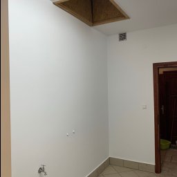 Szymon Lipka Usługi Budowlane - Wnętrze pomieszczenia z białymi ścianami, widoczne otwór w suficie z drewnianym obramowaniem, kran na ścianie, fragment drzwi i podłoga z płytek.