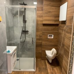 Szymon Lipka Usługi Budowlane - Wykończona łazienka z kabiną prysznicową typu walk-in z szarymi płytkami imitującymi marmur, drewnopodobnymi płytkami na ścianach i podłodze, białą szafką z umywalką oraz toaletą z grzejnikiem.