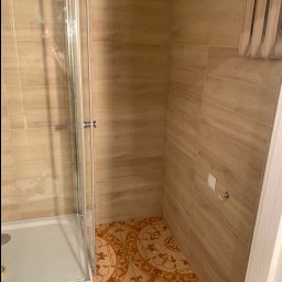Szymon Lipka Usługi Budowlane - Wykończona łazienka z kabiną prysznicową, brązowymi płytkami imitującymi drewno na ścianach i wzorzystą, brązowo-beżową podłogą. Widoczny fragment grzejnika.