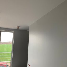 Szymon Lipka Usługi Budowlane - Wnętrze w szarościach po malowaniu: ściany, sufit i okno z widokiem na zieleń za oknem. Widoczne wystające kable elektryczne i niedokończone gniazdka na ścianie.