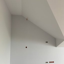 Szymon Lipka Usługi Budowlane - Wnętrze po malowaniu ścian i sufitu na jasny szary kolor, widoczne otwory na gniazdka elektryczne i wentylację, sufit skośny.