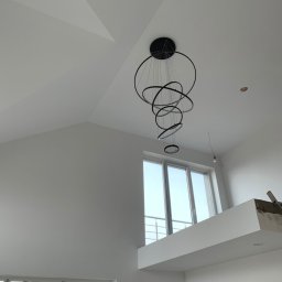 Szymon Lipka Usługi Budowlane - Wnętrze z wysokim sufitem i skośnymi ścianami, pomalowane na biało, z nowoczesną, okrągłą lampą wiszącą i dużymi oknami.