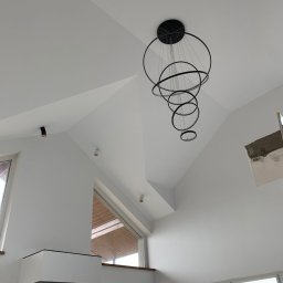 Szymon Lipka Usługi Budowlane - Wnętrze nowoczesnego domu z wysokim sufitem i skośnymi ścianami, pomalowanymi na biało, z widocznym czarnym, okrągłym żyrandolem i oknami z widokiem na zewnątrz.