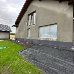 Szymon Lipka Usługi Budowlane - Montaż izolacji termicznej na elewacji domu jednorodzinnego, widoczne szare płyty izolacyjne przy gruncie, okna zabezpieczone folią, pracownik w trakcie pracy, teren zabezpieczony czarną folią.