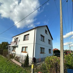 Szymon Lipka Usługi Budowlane - Dwupiętrowy dom z balkonem, świeżo pomalowaną białą elewacją, brązowymi ramami okiennymi i anteną satelitarną, widoczny z dołu pod kątem, z elementami rusztowania i drabiną opartą o ścianę, na tle...