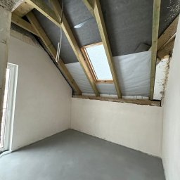 Szymon Lipka Usługi Budowlane - Poddasze z oknem dachowym w trakcie prac wykończeniowych: ściany pokryte gładzią, surowy drewniany szkielet dachu z widoczną membraną, wylewka na podłodze.