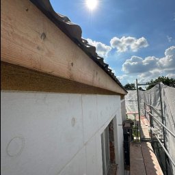 Szymon Lipka Usługi Budowlane - Ocieplanie elewacji budynku styropianem, widoczne rusztowanie, niebo z chmurami i słońce.