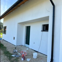 Szymon Lipka Usługi Budowlane - Świeżo otynkowana, biała elewacja domu jednorodzinnego z czarną rynną i stolarką okienną; na żwirowym podłożu widoczne narzędzia i pozostałości po pracach wykończeniowych.