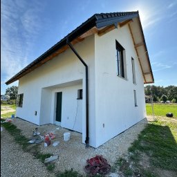 Szymon Lipka Usługi Budowlane - Świeżo otynkowany dom jednorodzinny z czarną stolarką okienną i dachem, widoczny z poziomu gruntu, z rozrzuconymi narzędziami i materiałami budowlanymi na żwirowym podłożu.