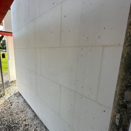 Szymon Lipka Usługi Budowlane - Ściana zewnętrzna budynku w trakcie ocieplania, widoczne białe bloki styropianowe z pionowymi rowkami, szara podstawa z kamieni, fragment rusztowania z prawej strony.