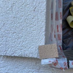 Szymon Lipka Usługi Budowlane - Fragment elewacji z próbką tynku o podobnej fakturze, zabezpieczony folią okienną z logo producenta.