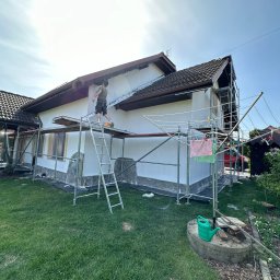 Szymon Lipka Usługi Budowlane - Pracownik na rusztowaniu aplikuje biały tynk na elewację domu jednorodzinnego w słoneczny dzień, widoczna suszarka na pranie i ogródek.