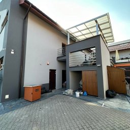 Szymon Lipka Usługi Budowlane - Dwukondygnacyjny dom z nową, dwukolorową elewacją w odcieniach szarości, balkonem z metalową balustradą i zadaszeniem nad garażem; na brukowanym podjeździe widoczne pozostałości po pracach...