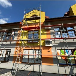 Szymon Lipka Usługi Budowlane - Budynek wielorodzinny w trakcie prac remontowych elewacji, widoczne rusztowania, fragmenty pomalowane na żółto, pomarańczowo i szaro, pracownik na rusztowaniu.