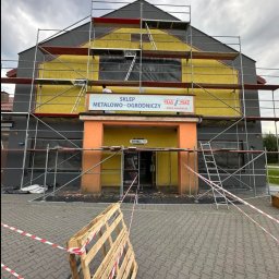 Szymon Lipka Usługi Budowlane - Sklep metalowo-ogrodniczy w trakcie prac elewacyjnych, widoczne rusztowania, częściowo pomalowana fasada na żółto i szaro, zabezpieczone okna folią, paleta na pierwszym planie.