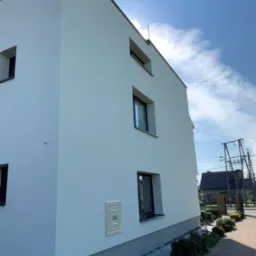 Nowoczesny, trzypiętrowy dom z białą elewacją i ciemnymi ramami okiennymi, widok z poziomu chodnika z kostki brukowej, obok skrzynka gazowa i skromna zieleń.