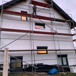 Szymon Lipka Usługi Budowlane - Pracownik na rusztowaniu, wykonuje prace na elewacji domu z białych bloczków, widoczne okna z roletami, oświetlone od wewnątrz, oraz zabezpieczenia gruntowe.