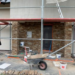 Szymon Lipka Usługi Budowlane - Montaż elewacji z kamienia dekoracyjnego na ścianie domu, widoczne rusztowanie, taczka, narzędzia i materiały budowlane, drzwi zabezpieczone folią.
