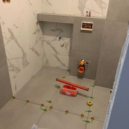 Szymon Lipka Usługi Budowlane - Łazienka w trakcie układania szarych płytek podłogowych i białych, marmurkowych płytek ściennych. Widoczne poziomica, miara, pojemniki z krzyżykami dystansowymi, rury i otwory instalacyjne.