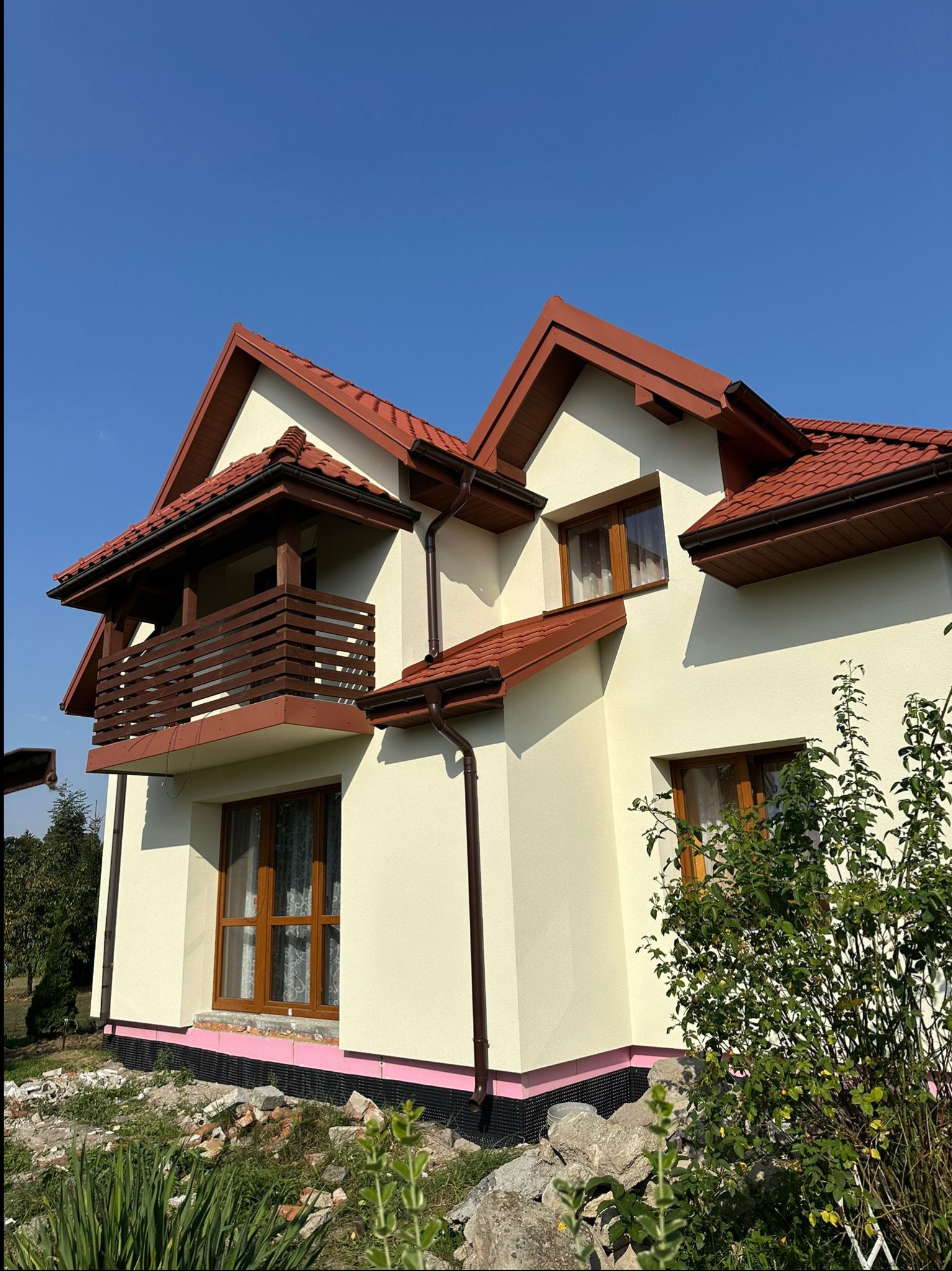 Nowy dom z beżową elewacją, brązowymi oknami i dachem z czerwonej dachówki, widok z dołu na balkon z drewnianą balustradą i ocieplenie fundamentów z różowego styropianu.