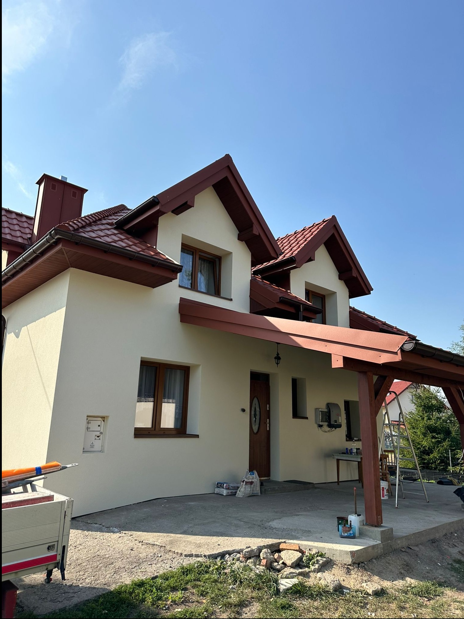 Jasna elewacja domu jednorodzinnego z brązową stolarką okienną i dachem, widoczny fragment przyczepy z narzędziami oraz materiały budowlane na podjeździe.