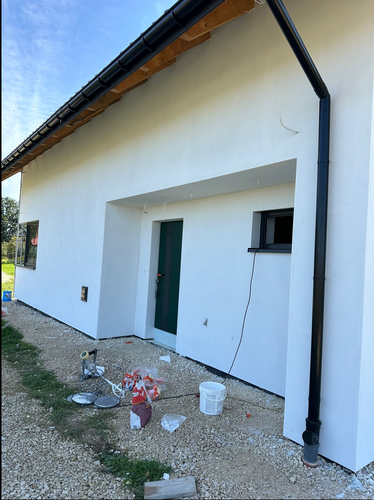 Świeżo otynkowana, biała elewacja domu jednorodzinnego z czarną rynną i stolarką okienną; na żwirowym podłożu widoczne narzędzia i pozostałości po pracach wykończeniowych.