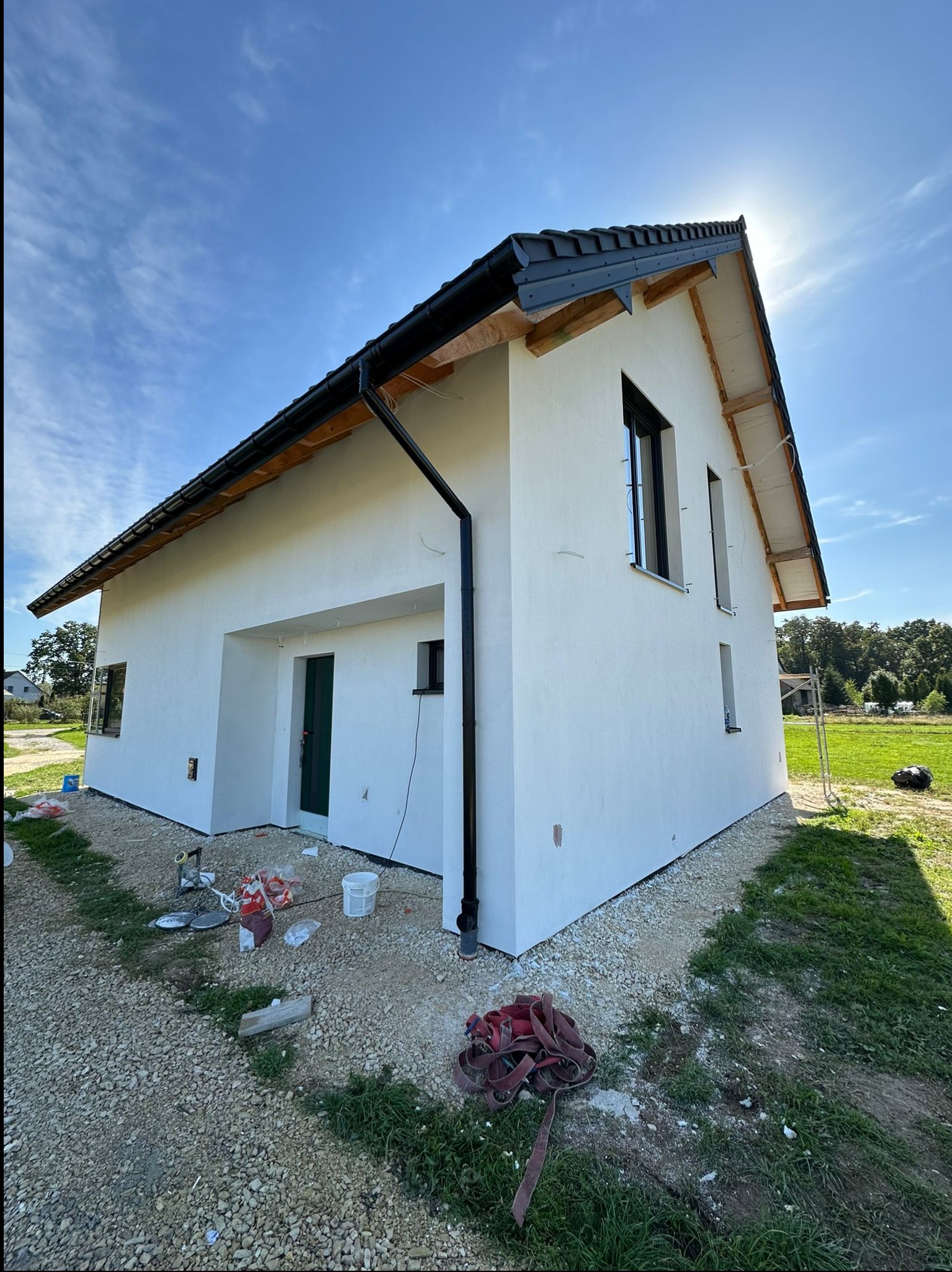 Świeżo otynkowany dom jednorodzinny z czarną stolarką okienną i dachem, widoczny z poziomu gruntu, z rozrzuconymi narzędziami i materiałami budowlanymi na żwirowym podłożu.
