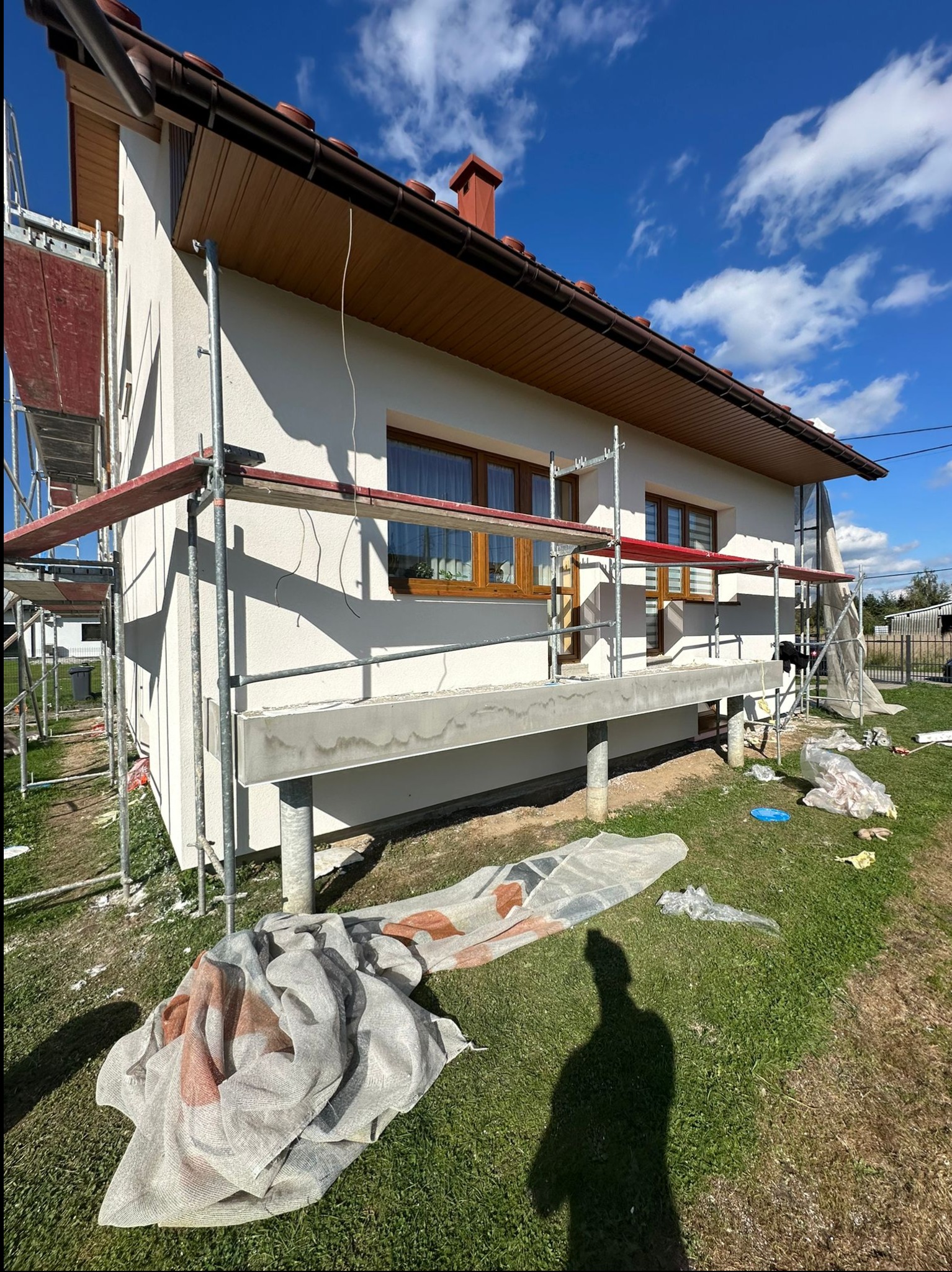 Dom jednorodzinny w trakcie prac elewacyjnych: biała ściana z brązowymi oknami, otoczona rusztowaniami, widoczne materiały budowlane i cień osoby na trawniku.