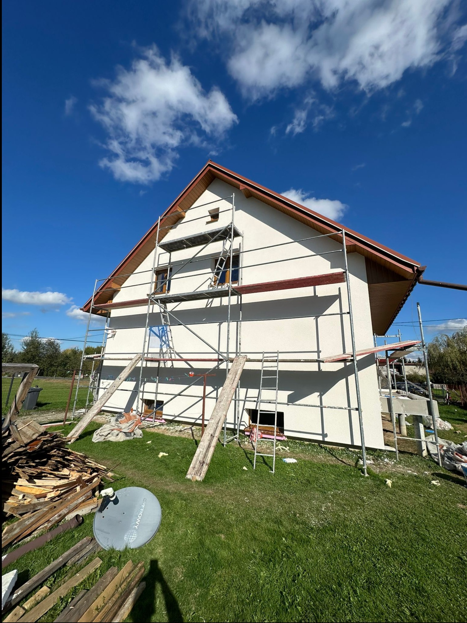 Dom jednorodzinny w trakcie prac elewacyjnych, widoczne rusztowania, świeżo otynkowana fasada w kolorze jasnego beżu, fragmenty desek i antena satelitarna na trawniku.