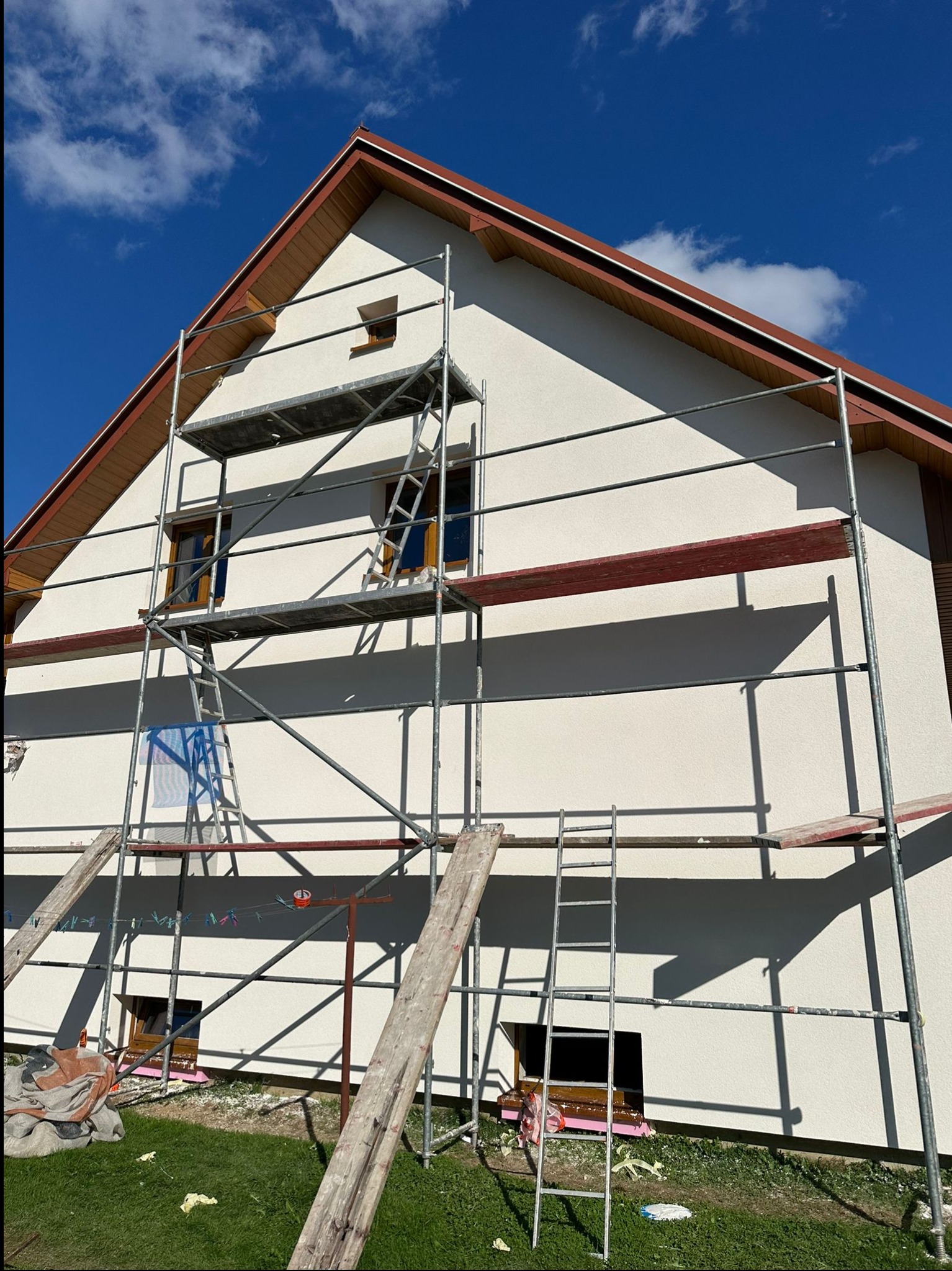 Dom jednorodzinny z jasną elewacją w trakcie prac wykończeniowych, widoczne rusztowania, fragment ocieplenia okien i błękitne niebo w tle.