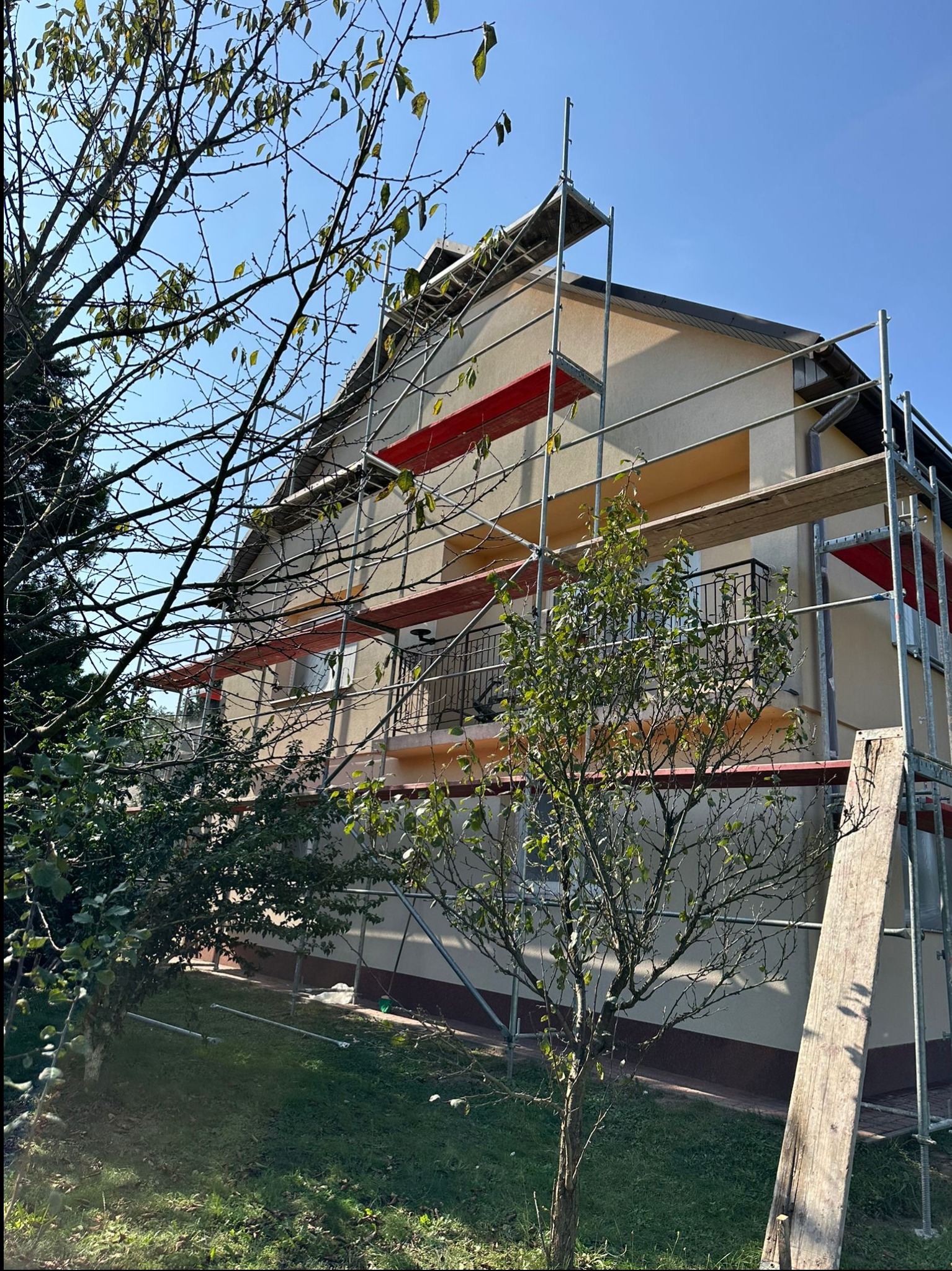 Dom jednorodzinny w trakcie prac elewacyjnych, widoczne rusztowania, fragment balkonu i drzewa na pierwszym planie. Ściany w jasnym odcieniu beżu.