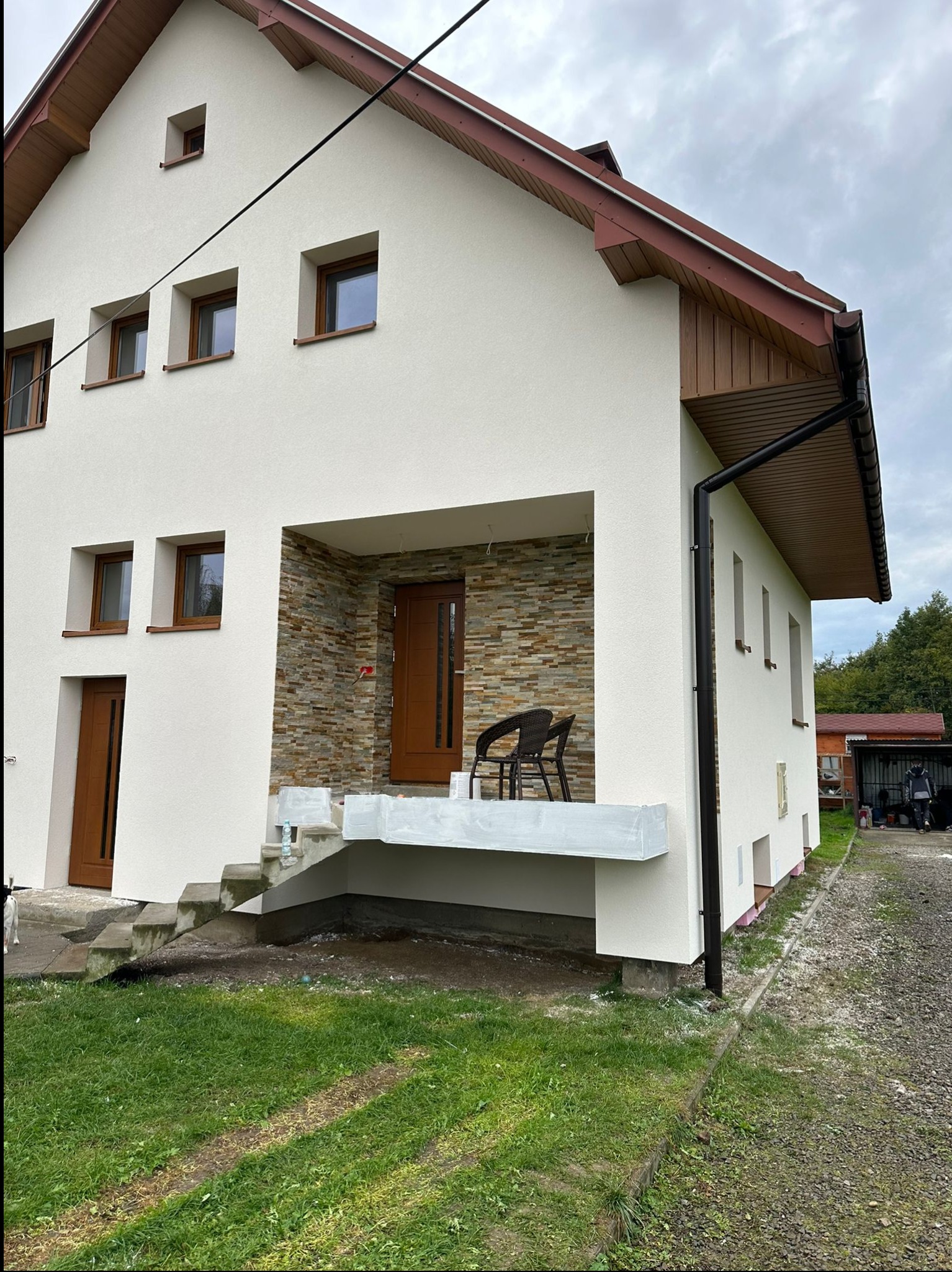 Dom jednorodzinny z nową, jasną elewacją i brązowymi ramami okiennymi, widoczne schody wejściowe z zabezpieczeniami malarskimi i fragment wykończonej ściany z kamienia dekoracyjnego.