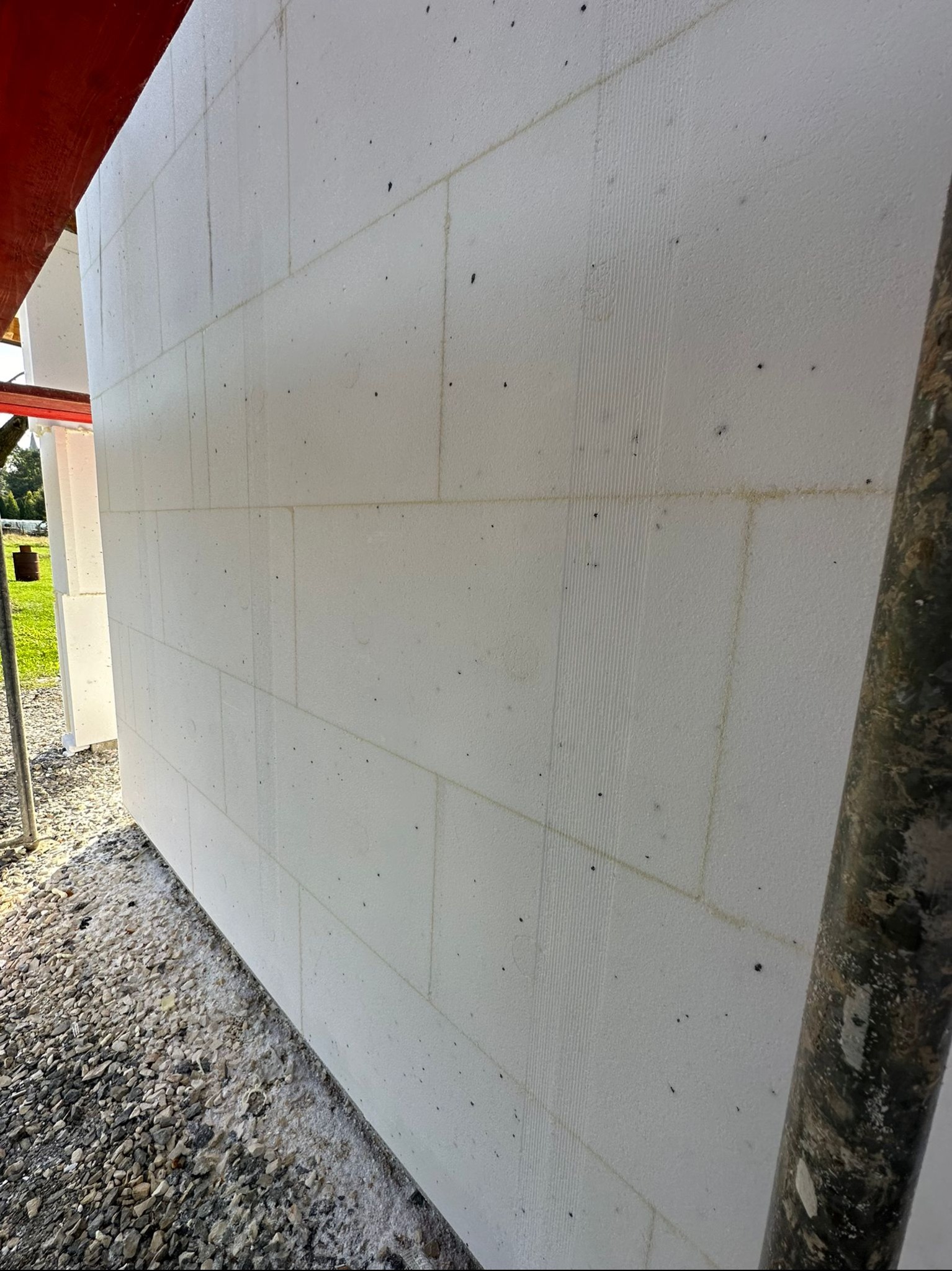 Ściana zewnętrzna budynku w trakcie ocieplania, widoczne białe bloki styropianowe z pionowymi rowkami, szara podstawa z kamieni, fragment rusztowania z prawej strony.