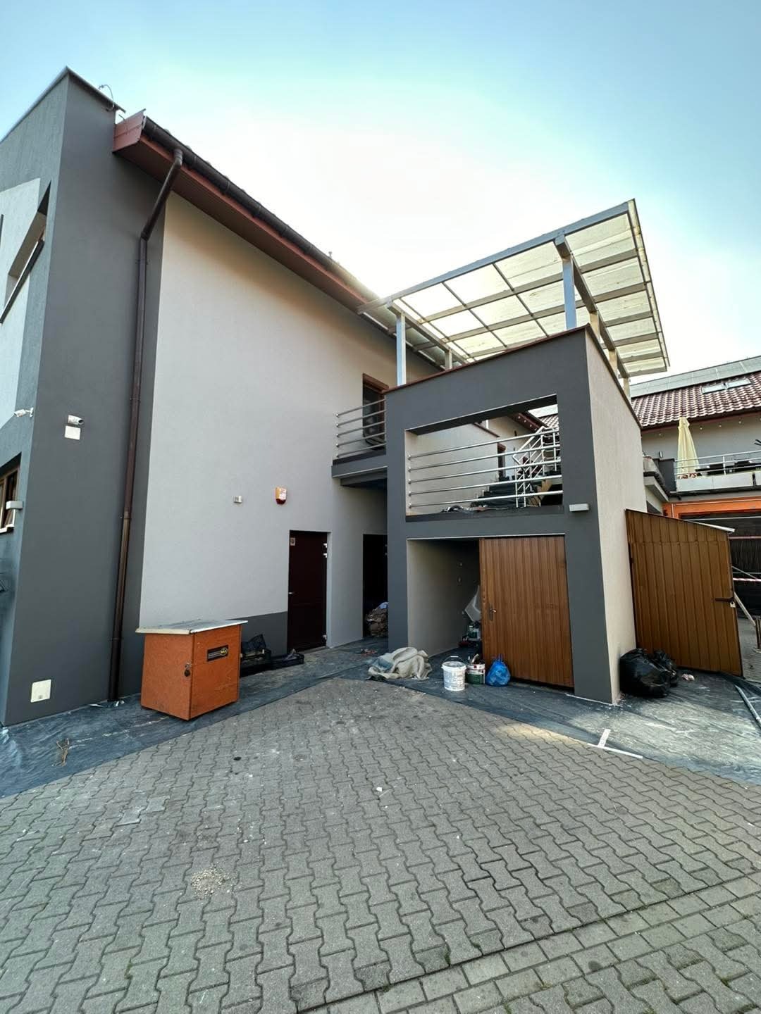 Dwukondygnacyjny dom z nową, dwukolorową elewacją w odcieniach szarości, balkonem z metalową balustradą i zadaszeniem nad garażem; na brukowanym podjeździe widoczne pozostałości po pracach...